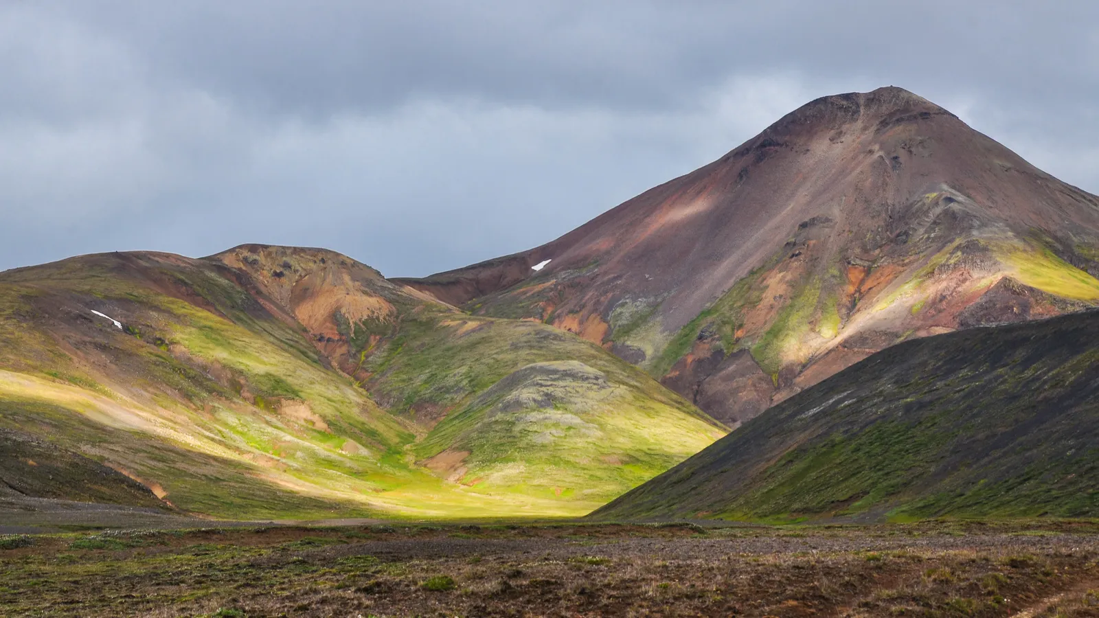 Green, ochre and reddish Highland slopes surrounding a sunlit valley beneath a cloudy sky
