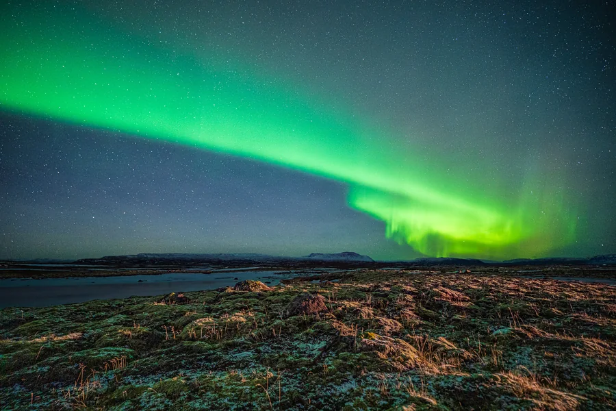 A green auroral arc across the sky above a rocky shoreline and open water