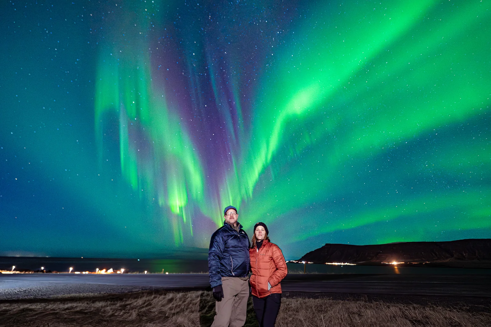 Two guests posing beneath green and purple northern lights near a shoreline