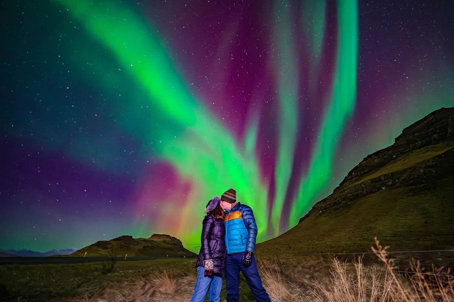 Two guests wearing warm hats, gloves and padded jackets beneath green and purple Northern Lights