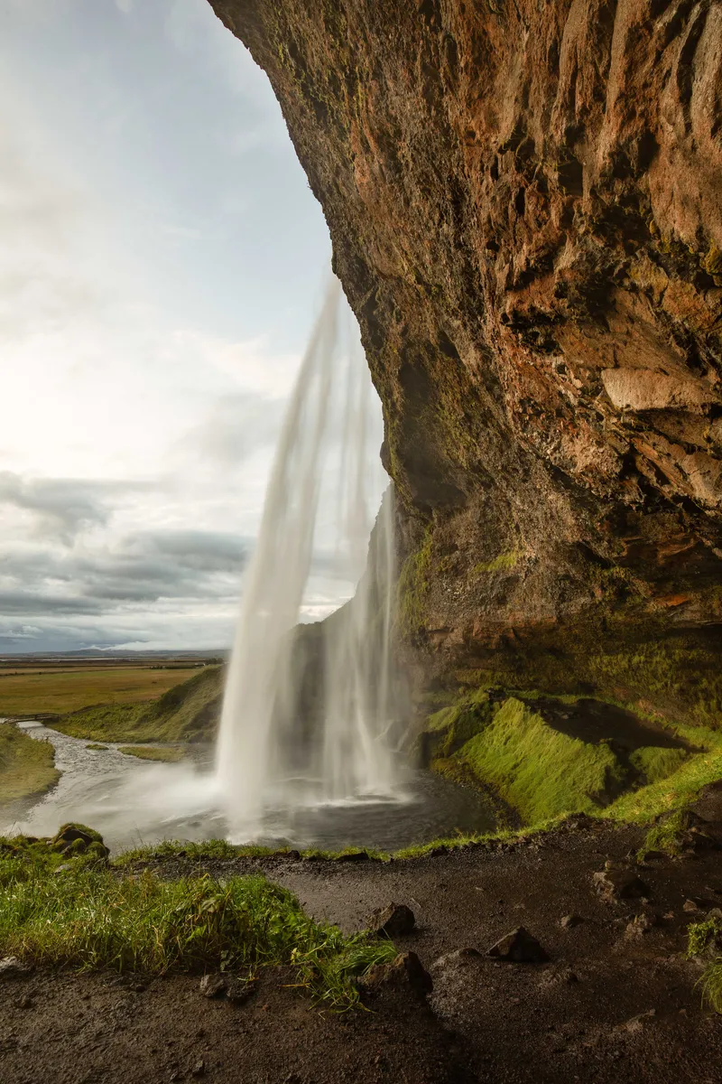 Seljalandsfoss seen from behind its falling water, with a rocky overhang and green fields beyond