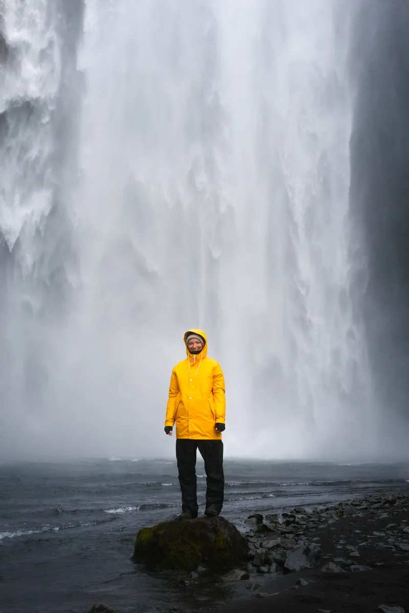A visitor in a yellow waterproof jacket standing below the falling water at Skógafoss
