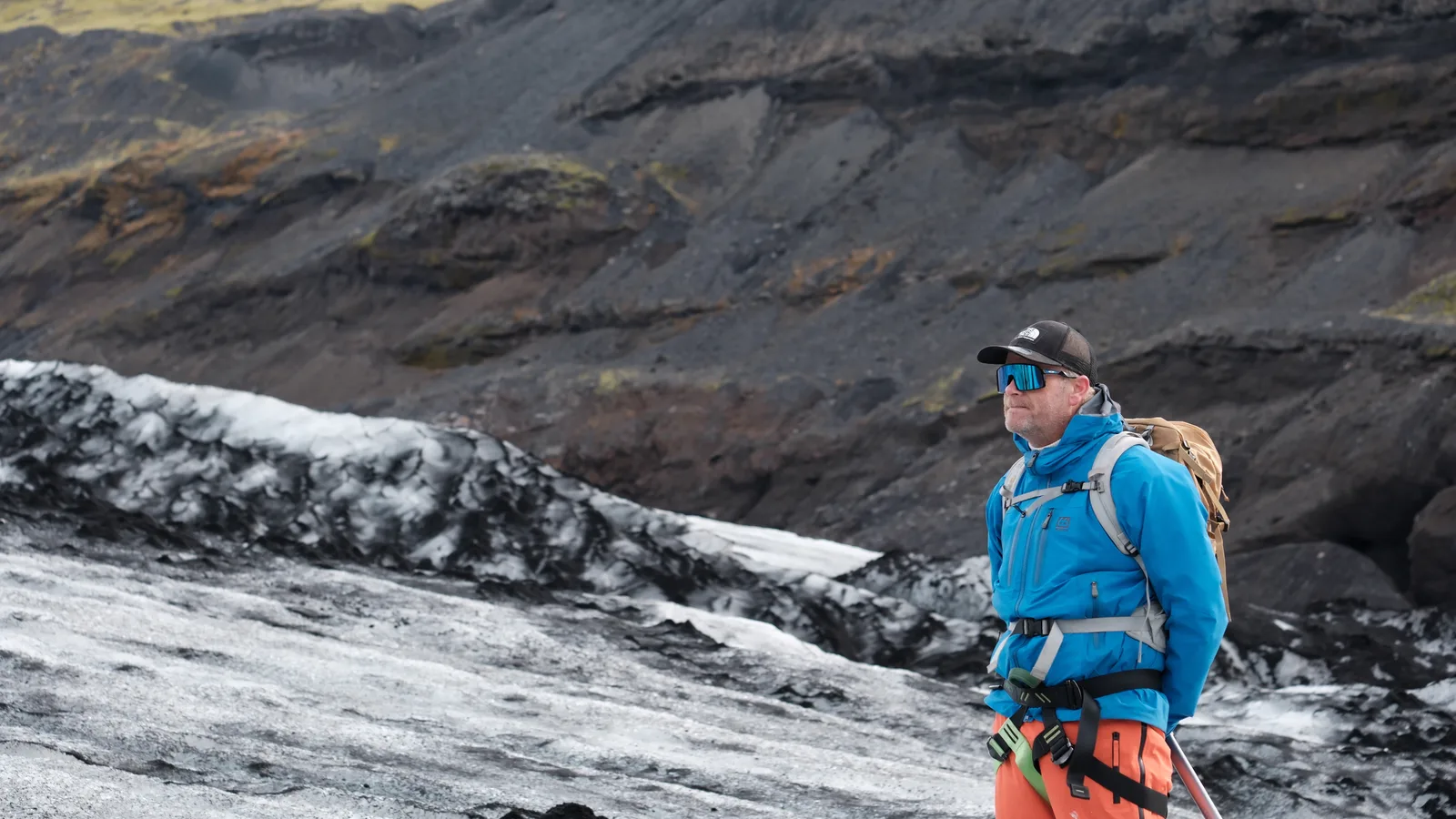 A hiker wearing a blue jacket, daypack and harness beside the ash-striped ice at Sólheimajökull