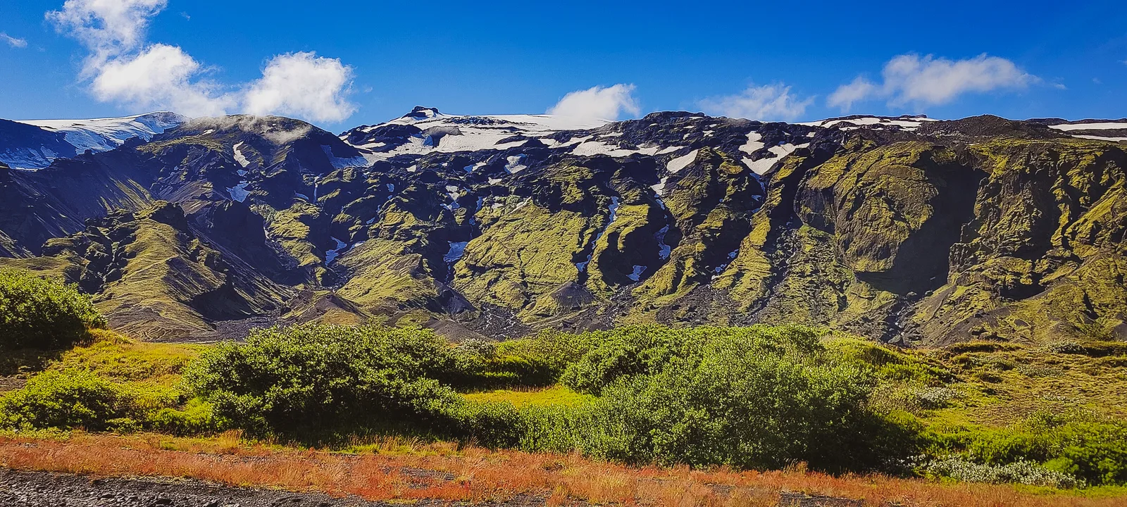 Green birch woodland beneath steep mountains and lingering snow in Þórsmörk