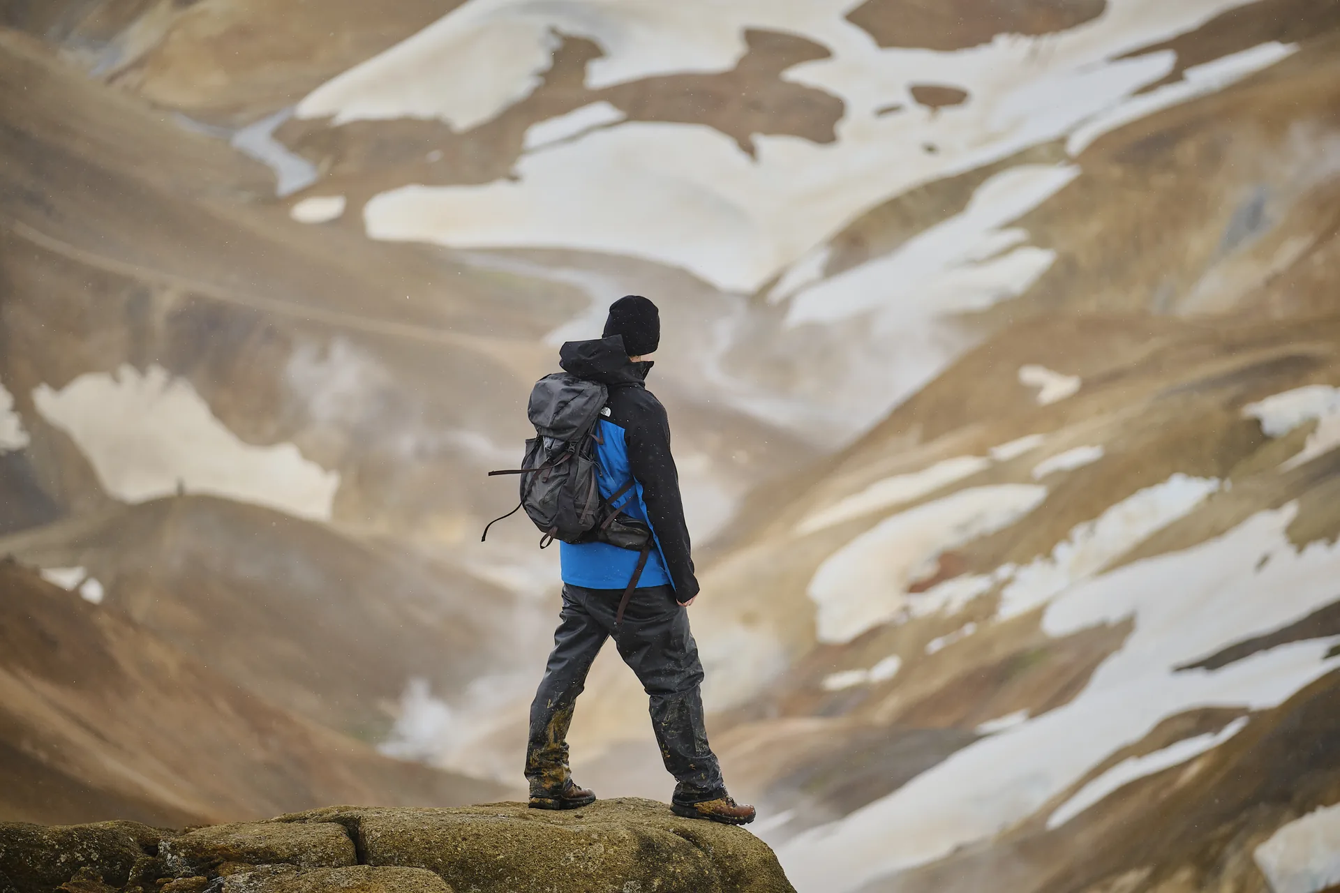 A hiker carrying a daypack and wearing a jacket and walking boots above snow-streaked Highland slopes